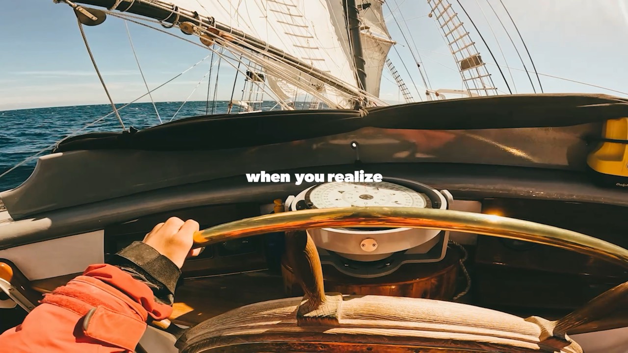 Point-of-view at the ship's wheel, a hand on the helm with the compass and sails ahead